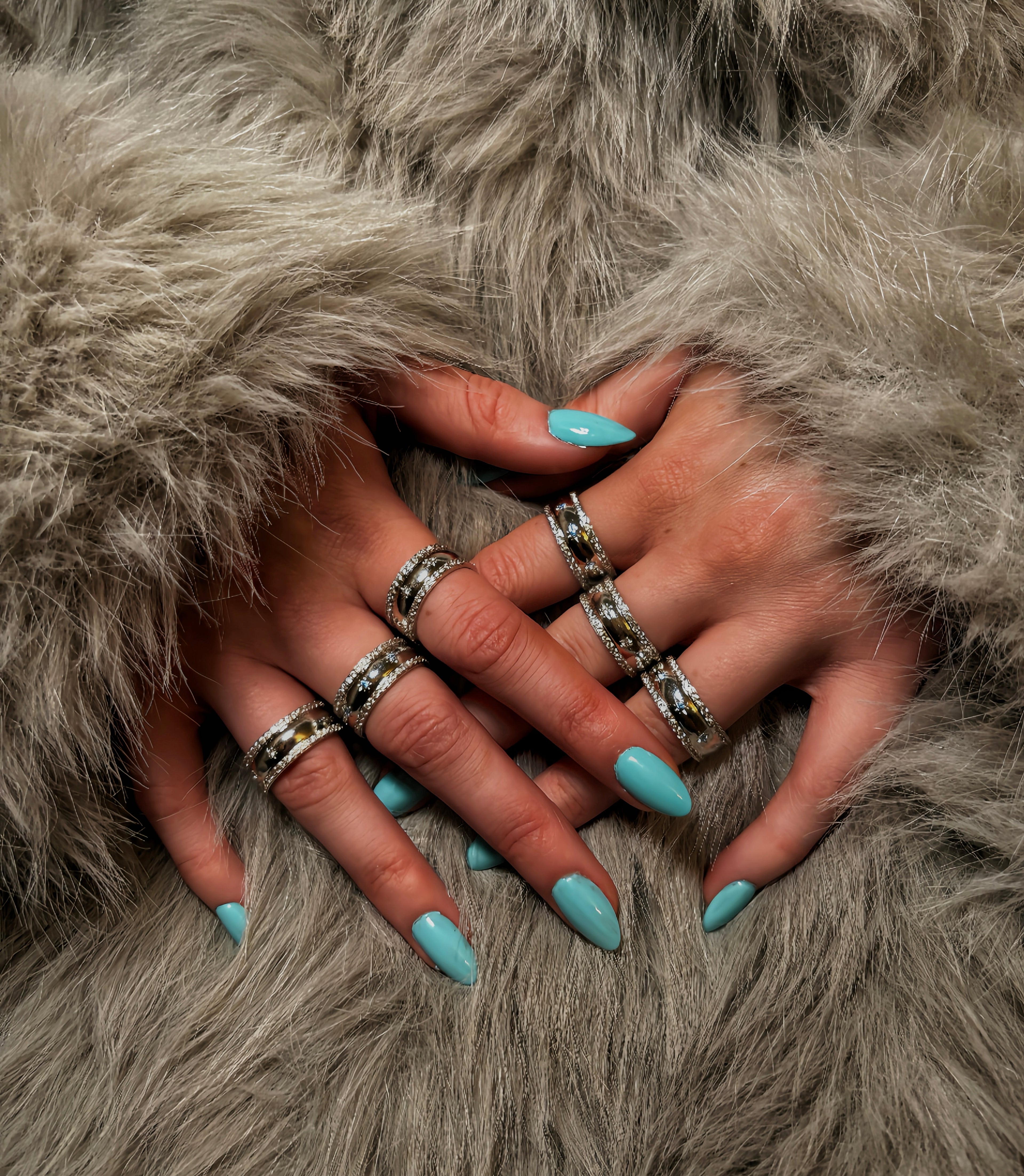 Close-up of hands with turquoise nail polish and multiple rings on a fur surface